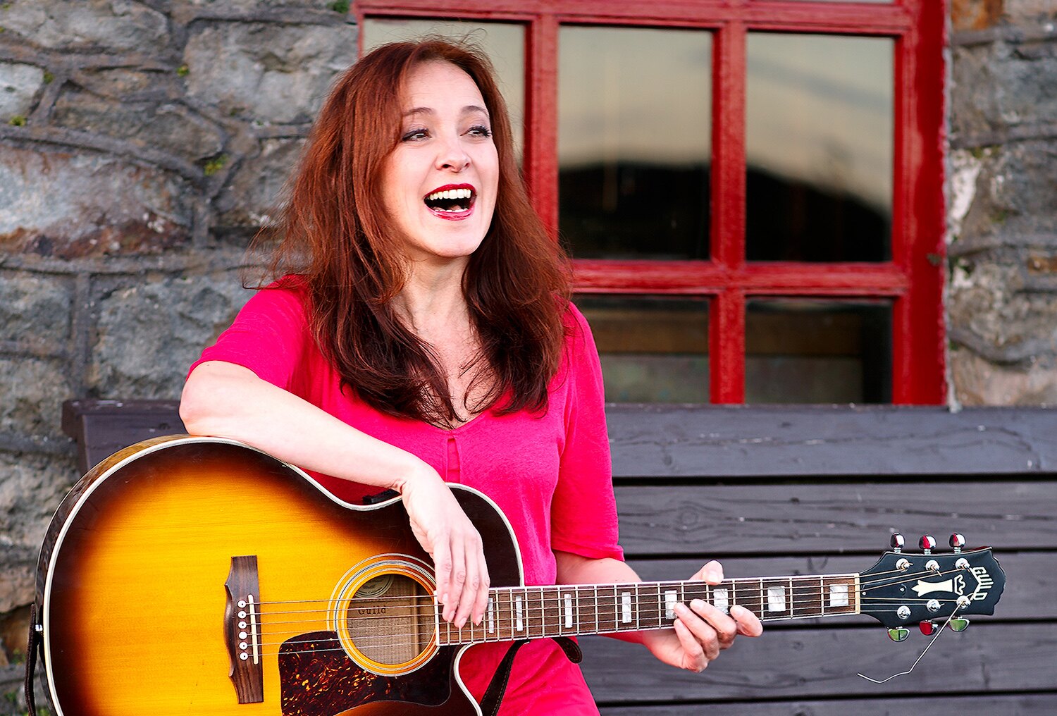 Gabriele Schuh lacht, im roten Kleid mit ihrer Gitarre vor einem roten Fenster in Irland